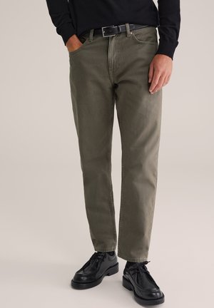 Mann trägt olivgrüne Hose mit geradem Bein, schwarzen Gürtel, schwarzes Langarmhemd und schwarze Schnürschuhe aus Leder, steht vor einfarbigem Hintergrund.