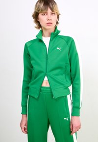 Green athletic zip-up jacket with white side stripes, made of smooth fabric. Features a high collar and front zipper, paired with matching pants.