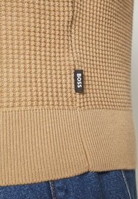 Beige gestrickter Pullover mit strukturiertem Muster, geripptem Saum und einem schwarzen Etikett mit der Aufschrift "BOSS" an der Seite. Kombiniert mit blauen Jeans.