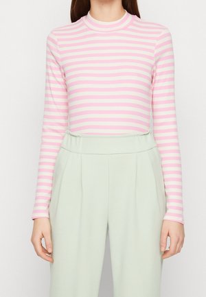 Mujer con una blusa de manga larga de rayas rosa y blancas metida en unos pantalones de pierna ancha de talle alto de color verde claro, posando contra un fondo blanco.