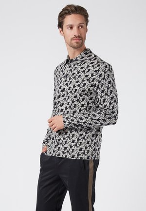 Camisa a rayas en blanco y negro con cuello, que presenta formas abstractas de hojas. Mangas largas, corte ajustado. Llevada con pantalones oscuros.