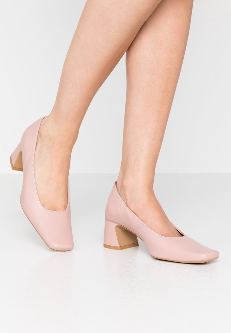 Femme portant des escarpins rose pâle à talon bas carré, debout sur un sol blanc devant un fond neutre.