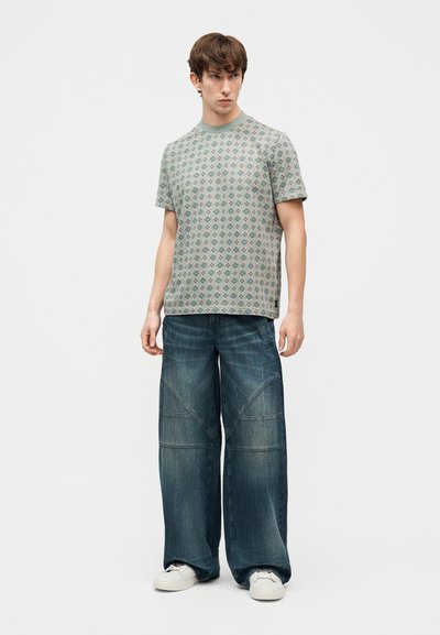 Νέος άνδρας στέκεται φορώντας κοντομάνικο μπλουζάκι με μοτίβο, φαρδιά ξεβαμμένα μπλε τζιν παντελόνια και λευκά αθλητικά παπούτσια, μπροστά σε απλό ανοιχτό φόντο.