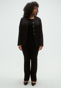 Fekete blézer ezüst gombokkal, testhez simuló szabás, vékony fekete nadrággal párosítva. A modell fekete felsőt és fényes fekete cipőt visel. Semleges háttér.