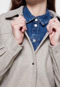 Chaqueta de lana gris claro con cierres de presión, superpuesta sobre una camisa azul de denim con tapeta de botones y cuello clásico.