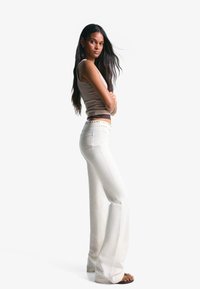 Bela razširjena jeans hlače z visokim pasom imajo okrasne zakovice na pasu. Model nosi bež prilegajočo majico na tanke naramnice.