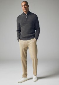 Maglione grigio scuro a maglia con colletto a mezza zip, abbinato a pantaloni beige e sneakers bianche, posando su uno sfondo neutro.