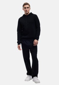 Czarna bluza z kapturem i sznurkami, teksturowany materiał, zestawiona ze luźnymi czarnymi spodniami i białymi sneakersami. Prosty design i neutralne kolory.