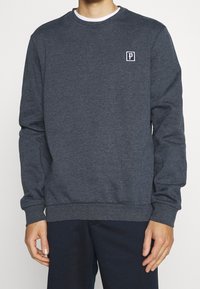 Uomo che indossa una felpa grigia scura a maniche lunghe con un piccolo logo bianco "P" sul petto sinistro e pantaloni scuri, in piedi contro uno sfondo neutro.