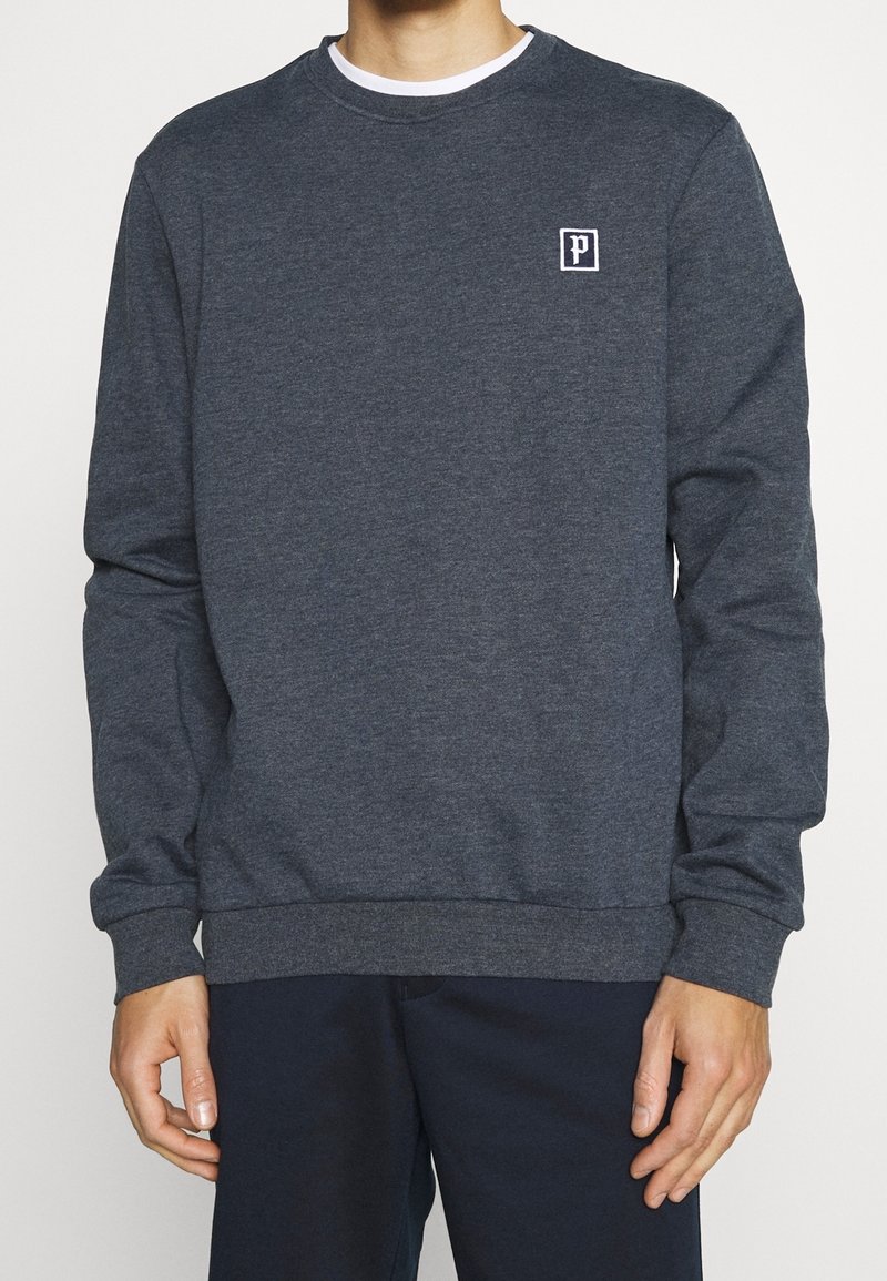 Uomo che indossa una felpa grigia scura a maniche lunghe con un piccolo logo bianco "P" sul petto sinistro e pantaloni scuri, in piedi contro uno sfondo neutro.