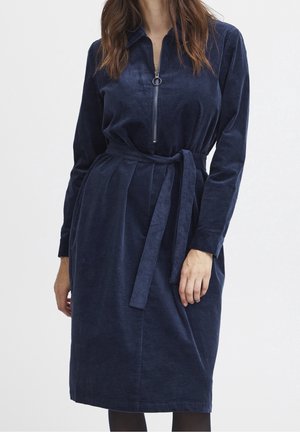 Vrouw draagt een marineblauwe corduroy jurk met lange mouwen, een rits aan de voorkant en een bijpassende stoffen riem gestrikt in de taille.