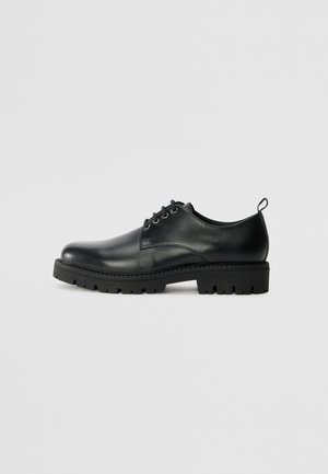 Zwarte lederen schoen met een ronde neus, voorzien van een vetersluiting, stevige zwarte rubberen zool en een klein hieltabje aan de achterkant.