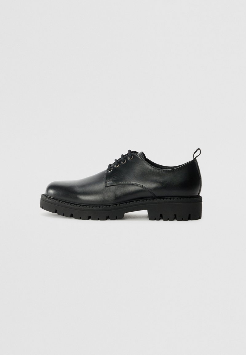 Zapato de cuero negro con punta redonda, que presenta un diseño de cordones, suela negra de goma resistente y una pequeña lengüeta en el talón.
