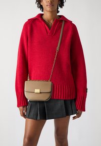 Maglione rosso a maglia con collo a V, abbinato a una gonna a pieghe a quadri nera. Una piccola borsa a tracolla color tan con una chiusura dorata.