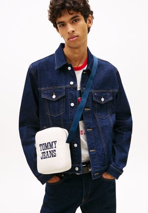 Ung mand iført en mørk denimjakke og jeans, hvid Tommy Jeans crossbody-taske, med hænderne i lommerne mod en ensfarvet baggrund.