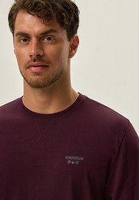 Burgundowa, długorękawowa koszula typu crewneck z bawełny, z małym szarym logo na klatce piersiowej i prostą, gładką fakturą.