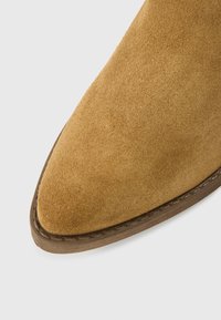 Bota de cano curto em camurça cor castanho claro, com bico pontiagudo, costura subtil ao longo da sola e textura suave. Destaca-se pela sola plana de borracha castanha.