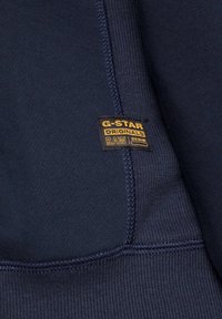 Tissu bleu marine foncé avec un ourlet cousu et une petite étiquette de marque G-Star Originals RAW Denim noire et jaune.