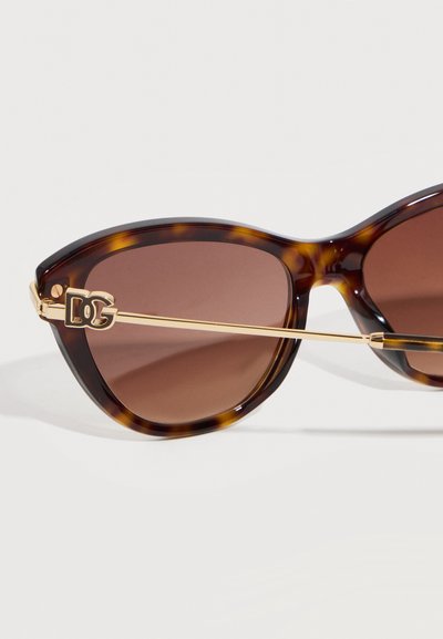 Γυαλιά ηλίου με καφέ φακούς σε σχέδιο γατόπαρδου, με λεπτούς χρυσούς βραχίονες και διακοσμητικό χρυσό λογότυπο κοντά στις μεντεσέδες.