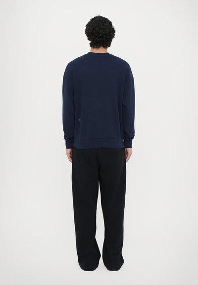 Mørkeblå sweatshirt med rund halsudskæring, ribbede manchetter og kant; parret med løstsiddende sorte bukser. Blødt, bomuldsagtigt materiale.