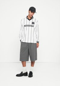 Chemise blanche à manches longues avec des rayures verticales noires et un col en V noir, associée à un short gris jusqu'aux genoux et des mocassins noirs.