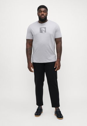 Svijetlosiva majica kratkih rukava s crno-bijelim grafičkim motivom lubanje. Uparena s crnim hlačama i tamnim tenisicama.