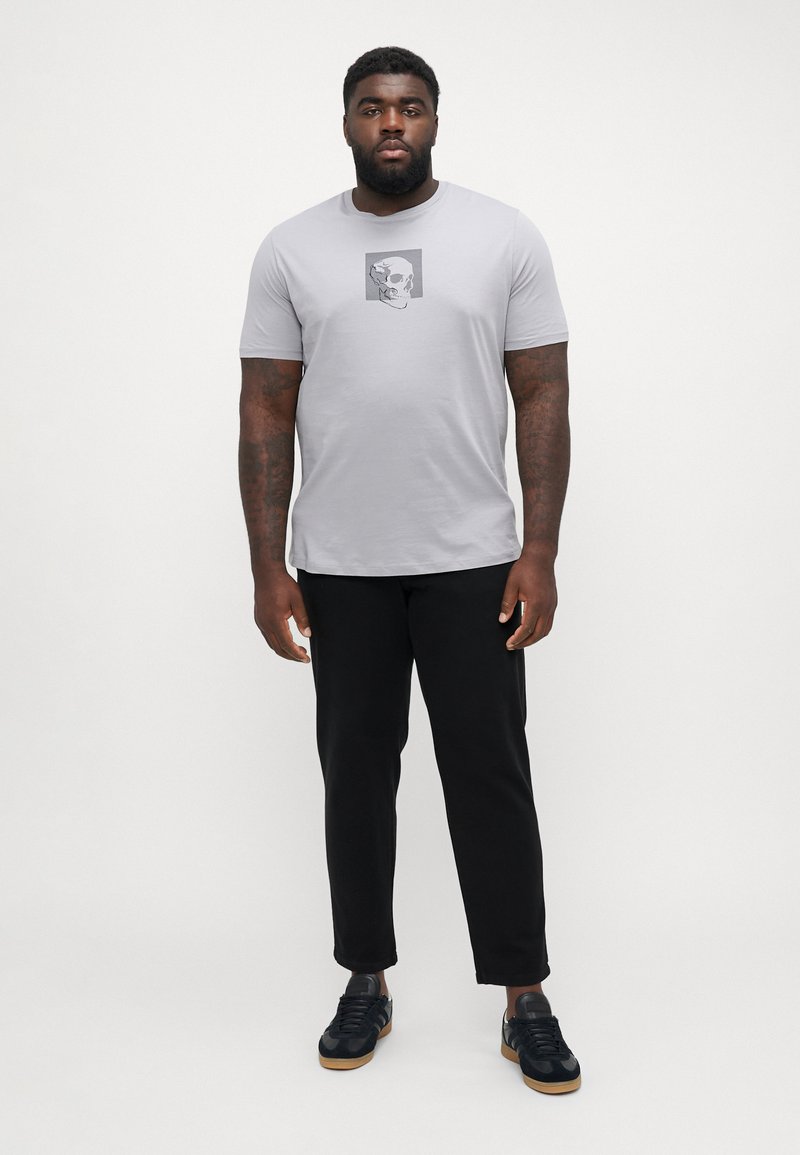 Hele halli lühikeste varrukatega t-särk, millel on must-valge kolju graafika. Kombineeritud mustade pükste ja tumedate spordijalatsitega.