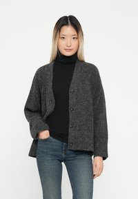 Kardigan in Anthrazitgrau mit entspannter Passform, versehen mit einem Knopfverschluss, weiten Ärmeln und weichem, strukturiertem Stoff. Trägt man über einem schwarzen Rollkragenpullover.