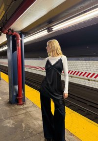 Fekete selyem hálóing fehér hosszú ujjú póló alatt, széles lábú fekete nadrággal. Háttérben egy metróállomás sárga peronnal.