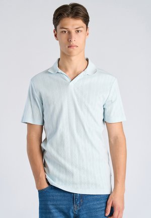 Giovane uomo con una polo a maniche corte azzurra con motivo e jeans blu, in piedi con una mano in tasca contro uno sfondo bianco.