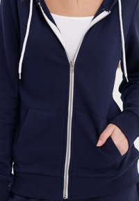 Sudadera con cremallera color navy hecha de una tela suave. Cuenta con una cremallera frontal, cordones blancos y bolsillos laterales con puños y dobladillo acanalados.