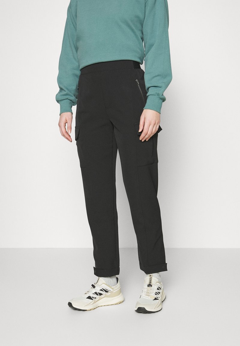 Pantalons cargo noirs avec taille élastique, poches latérales et poignets aux chevilles, associés à un sweat-shirt teal et des baskets beiges.
