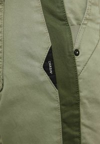 I pantaloni in cotone verde presentano un accento di tasca nera a contrasto con dettagli di cucitura. Il tessuto ha una texture liscia e un design snello.