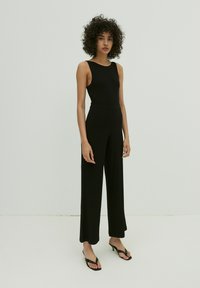 Schwarzer gerippter Jumpsuit mit tailliertem Oberteil, weitem Bein und ärmellosem Design. Getragen mit schwarzen Flip-Flops, vor einem schlichten Hintergrund.