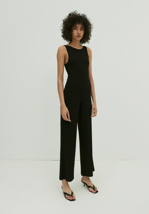 Schwarzer gerippter Jumpsuit mit tailliertem Oberteil, weitem Bein und ärmellosem Design. Getragen mit schwarzen Flip-Flops, vor einem schlichten Hintergrund.