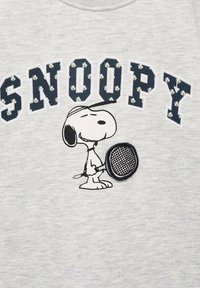 Snoopy drži teniški lopar, se smeška z zaprtimi očmi in nosi kapo; nad njim je napis "SNOOPY" z majhnimi belimi cvetlicami na črkah.