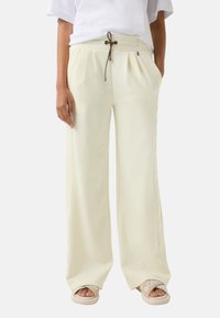 Pantalons larges en crème clair avec des plis avant, dotés d'un cordon de serrage marron et de poches latérales, associés à des sandales à glisser beiges.