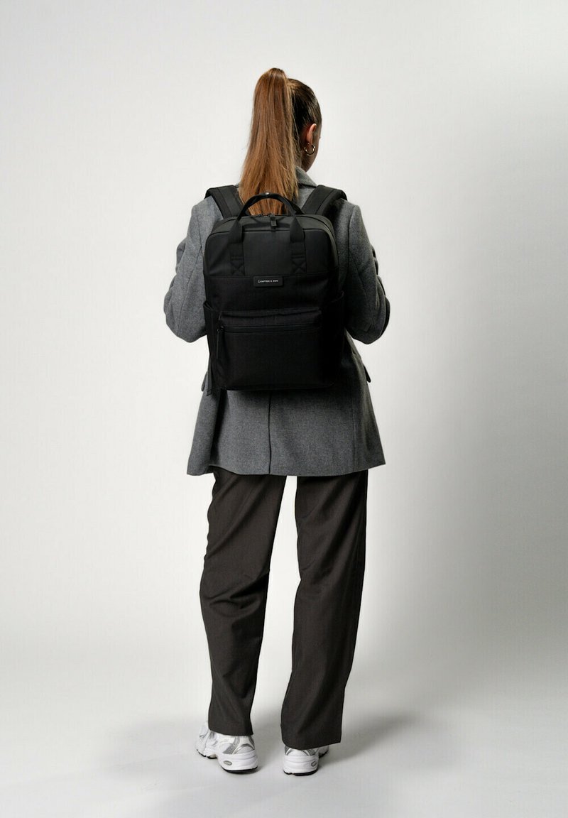 Zaino in tessuto nero con due spallacci spessi, una tasca frontale con zip e forma rettangolare. Indossato da una figura con un cappotto grigio e pantaloni marroni.