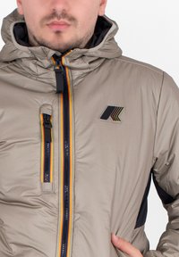 Giacca imbottita beige con cappuccio. Presenta una cerniera colorata con strisce rosse, gialle e blu navy. Toppa con logo sul petto. Tessuto strutturato.