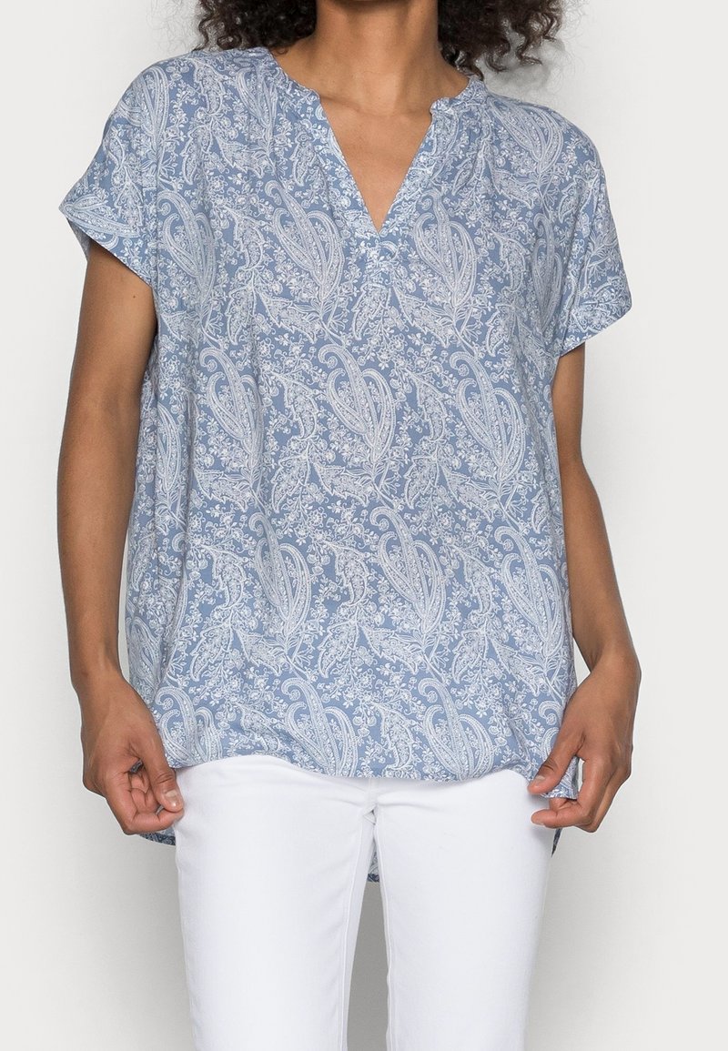 Femme portant un chemisier à manches courtes bleu clair avec motif cachemire blanc et un pantalon blanc, debout devant un fond uni.