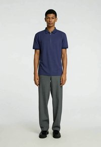 Ung mann står, iført en marineblå kortermet polo-skjorte, grå vide bukser og svarte sko på en ensfarget bakgrunn.