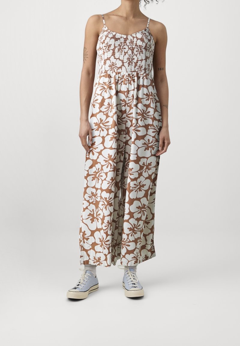 Personne portant une combinaison sans manches marron et blanche à fleurs, associée à des chaussettes blanches et des baskets bleu clair, debout devant un fond uni.