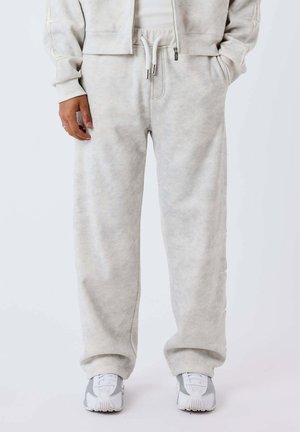 Personne portant un pantalon de jogging gris clair avec taille à cordon et baskets gris clair assorties, debout devant un fond uni.