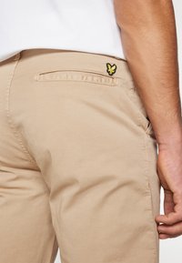 Beige Hose mit einem kleinen gestickten gelb-schwarzen Schmetterlingslogo über der Gesäßtasche, kombiniert mit einem weißen Hemd.