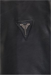 Schwarzes Leder mit strukturiertem Finish, ausgestattet mit einem dreieckigen Emblem mit einem metallischen Abzeichen in der Mitte.