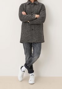 Abrigo gris texturizado con cierre de botones en el frente, dos bolsillos y cuello, combinado con jeans azules desgastados y zapatillas blancas.