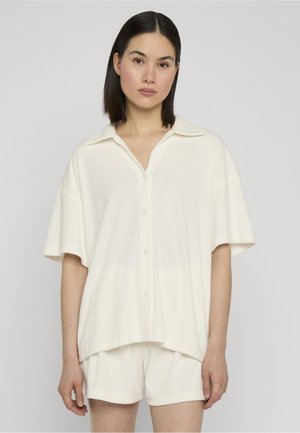 Frau mit glattem dunklem Haar, die ein lockeres weißes kurzärmeliges Hemd mit Knöpfen und passende weiße Shorts vor einem schlichten Hintergrund trägt.