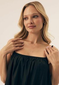 Czarny bezrękawnik z marszczonym materiałem przy dekolcie, miękka tekstura i regulowane ramiączka; modelka dotyka ramion, pokazując gładką skórę.