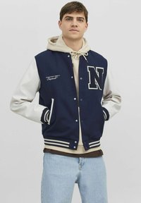 Jonge man in een marineblauwe en witte varsityjack met een 'N'-patch, een beige hoodie en lichtblauwe jeans, met zijn handen in de zakken van het jack.