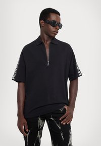 Melna polo krekla ar rāvējslēdzēju, īsām piedurknēm, kurās ir balta Versace logotipu lente. Valkāts ar melniem džinsiem ar izbalējušu rakstu.